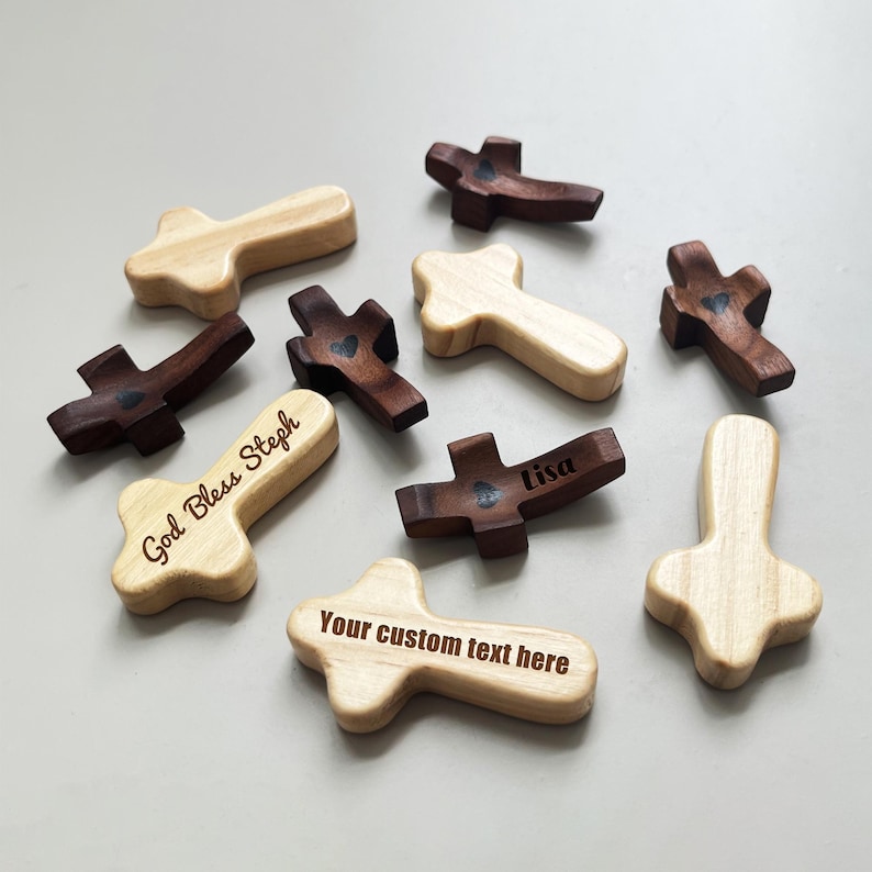 May include: A collection of wooden crosses in various shades of brown and natural wood. Some crosses have engraved text, including "God Bless Steph", "Lisa", and "Your custom text here". Several crosses feature a small heart design.
