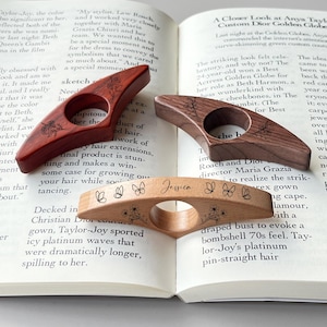 May include: Three wooden thumb page holders rest on an open book. One is dark red, another is brown, and the third is light wood with the name "Jessica" and butterfly and floral designs.