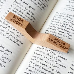 Puede incluir: Un sujetap&aacute;ginas de madera con las palabras "BOOK WHORE" y "SPREAD THOSE PAGES" grabadas. El soporte curvo descansa sobre las p&aacute;ginas abiertas de un libro, con texto visible.