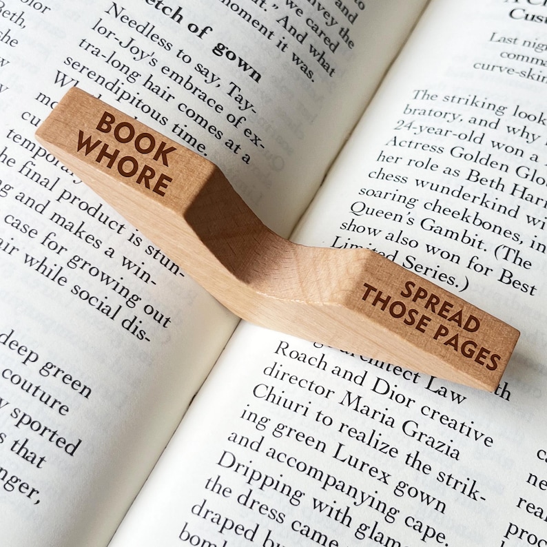 Puede incluir: Un sujetap&aacute;ginas de madera con las palabras "BOOK WHORE" y "SPREAD THOSE PAGES" grabadas. El soporte curvo descansa sobre las p&aacute;ginas abiertas de un libro, con texto visible.