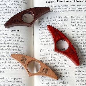 May include: Three wooden thumb page holders in varying shades of brown and red, each with a circular opening. Two have etched floral designs, while the third features a tree design. They rest on an open book.
