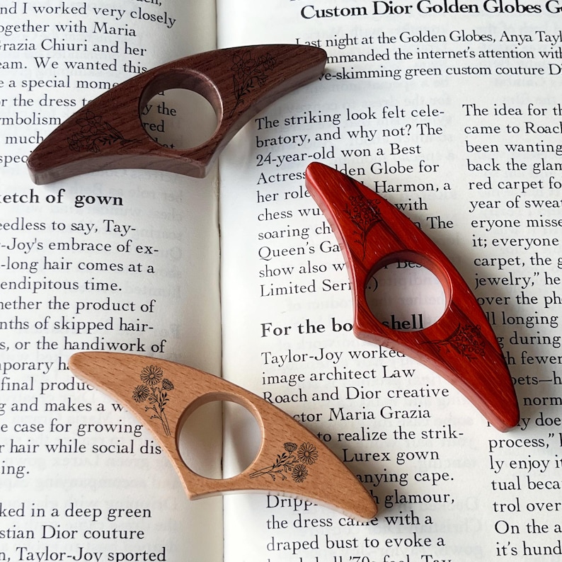 May include: Three wooden thumb page holders in varying shades of brown and red, each with a circular opening. Two have etched floral designs, while the third features a tree design. They rest on an open book.