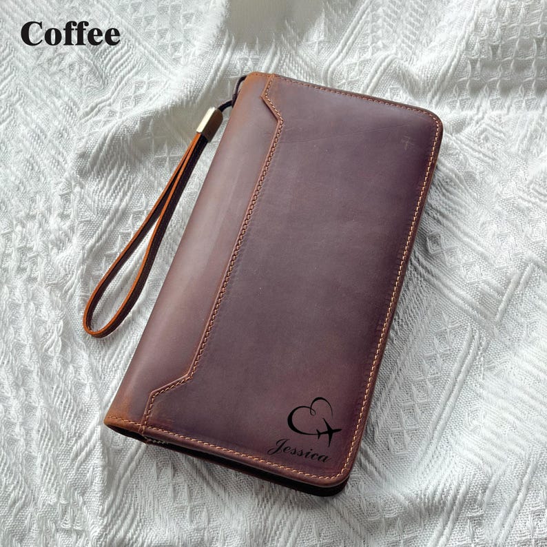May include: A brown leather wallet with a wrist strap. The wallet has a stitched design and the name "Jessica" is printed on the front, along with a heart and airplane graphic. The word "Coffee" is in the top left corner.