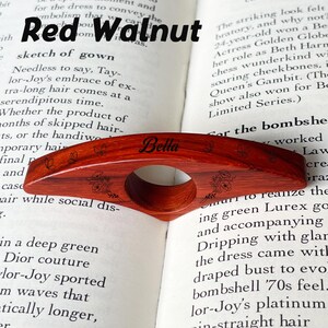 May include: A red walnut wood book page holder with the name "Bella" engraved on it. The holder has a circular opening for the finger and is decorated with floral and butterfly designs. The text "Red Walnut" is at the top.