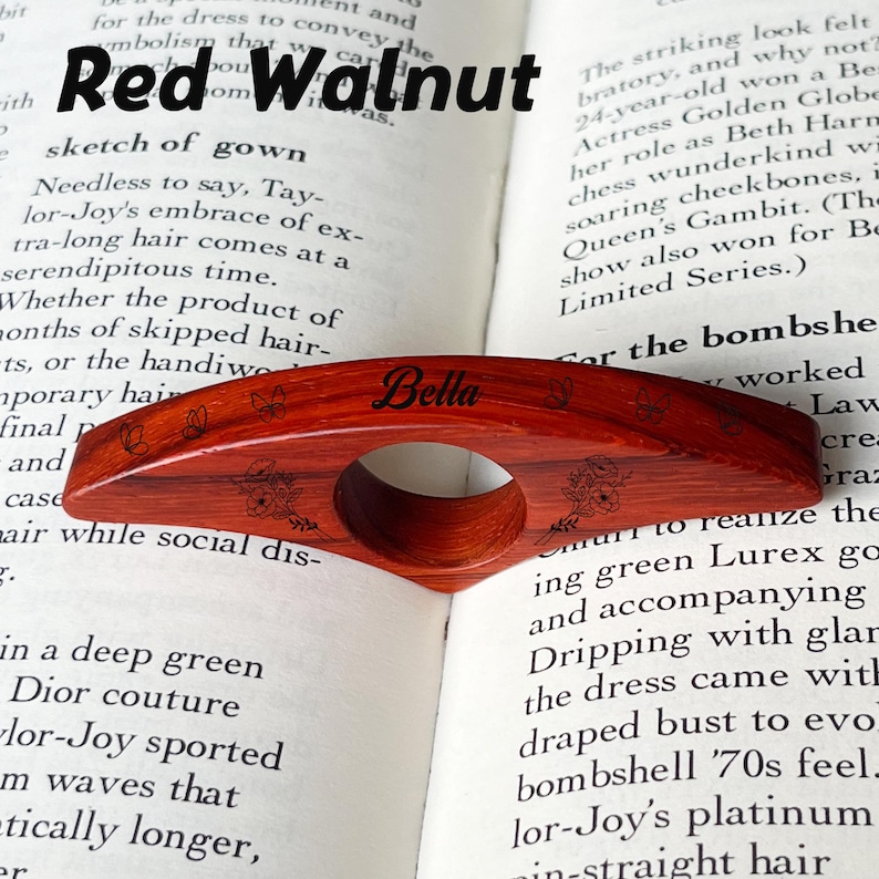 May include: A red walnut wood book page holder with the name "Bella" engraved on it. The holder has a circular opening for the finger and is decorated with floral and butterfly designs. The text "Red Walnut" is at the top.