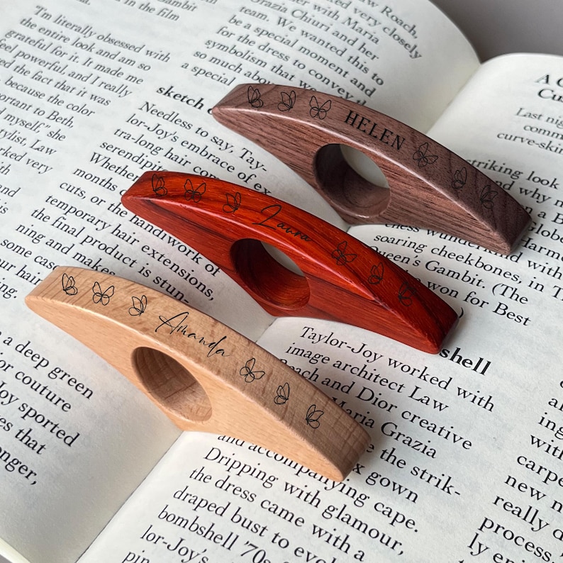 May include: Three wooden thumb page holders with butterfly designs and personalized names. The holders are in shades of brown, red, and light wood, resting on the pages of an open book.