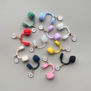 May include: A collection of colorful headphone-shaped charms in various colors, including green, blue, red, white, yellow, navy, pink, and black. Each charm has a gold-colored chain and a small, white circular tag with a letter.