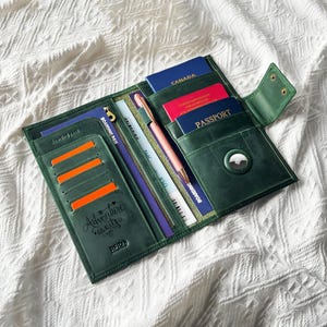 Peut inclure: Portefeuille de voyage en cuir vert ouvert, avec plusieurs compartiments. Le portefeuille contient un passeport, des cartes d'embarquement et un stylo. Le texte "Adventure awaits" est visible sur le devant. Le portefeuille est ouvert sur une surface blanche texturée.