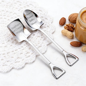 Puede incluir: Dos cucharas de acero inoxidable en forma de pala. Una cuchara está grabada con "Sophia's Ice Cream Shovel", y la otra con "Olivia's Peanut Butter Shovel". Las cucharas están sobre un mantel individual de encaje blanco, con nueces y un tarro de mantequilla de cacahuete en el fondo.