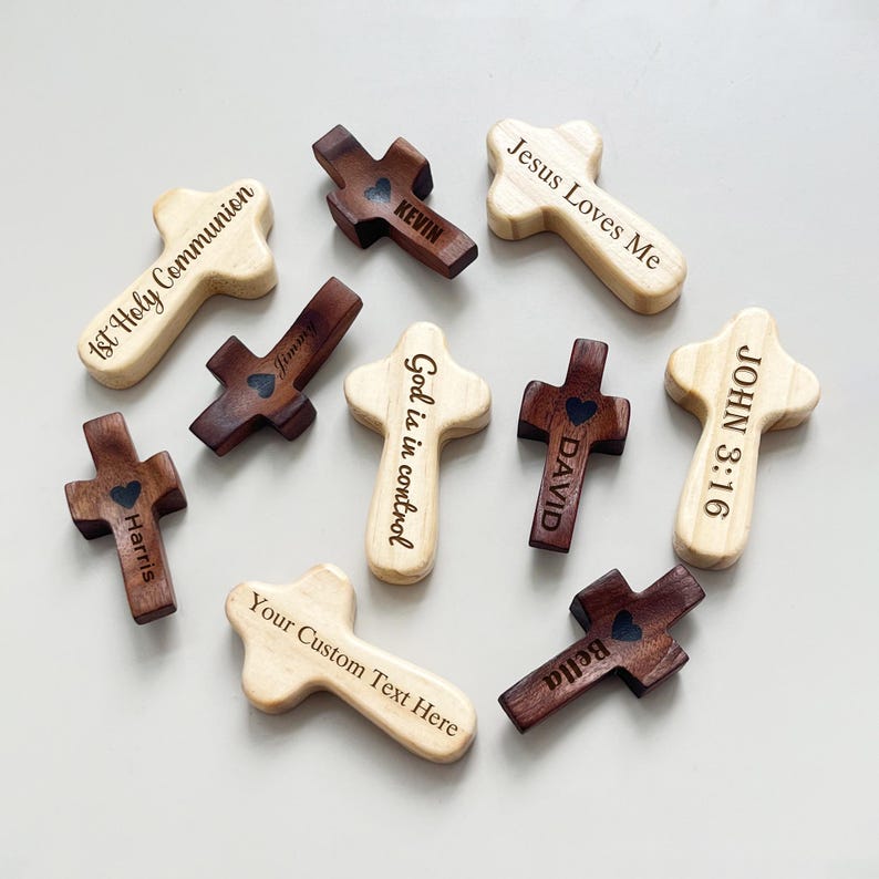May include: A collection of wooden crosses in various shades of brown and beige. Some crosses have engraved text such as "Jesus Loves Me," "God is in control," and names like "Kevin" and "David." Each cross features a heart detail.