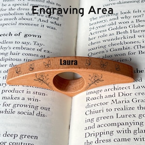 May include: A wooden book page holder with the name "Laura" engraved on it. The holder features a curved design with floral and butterfly engravings. It is resting on the pages of an open book.