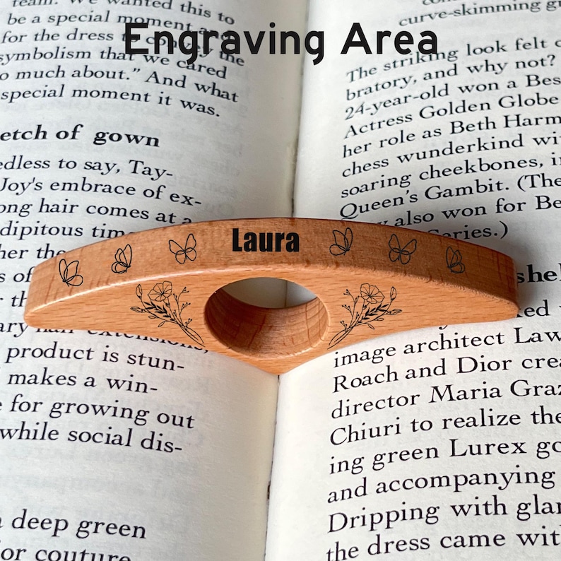 May include: A wooden book page holder with the name "Laura" engraved on it. The holder features a curved design with floral and butterfly engravings. It is resting on the pages of an open book.
