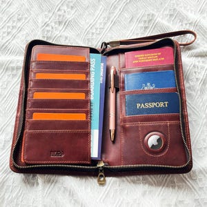 May include: Open brown leather travel wallet with multiple card slots, a boarding pass, and a pen. The wallet contains a passport and other documents. A wrist strap and zipper closure are visible.