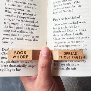 Puede incluir: Marcap&aacute;ginas de madera con las palabras "BOOK WHORE" y "SPREAD THOSE PAGES" impresas. El marcap&aacute;ginas est&aacute; colocado entre las p&aacute;ginas de un libro, con el texto visible.