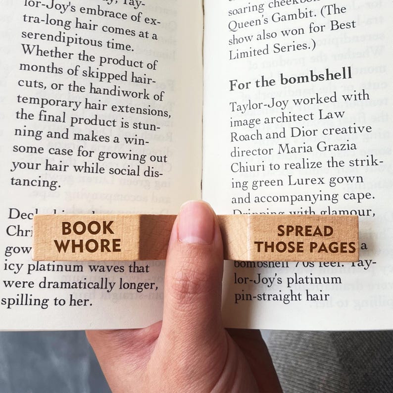 Puede incluir: Marcap&aacute;ginas de madera con las palabras "BOOK WHORE" y "SPREAD THOSE PAGES" impresas. El marcap&aacute;ginas est&aacute; colocado entre las p&aacute;ginas de un libro, con el texto visible.