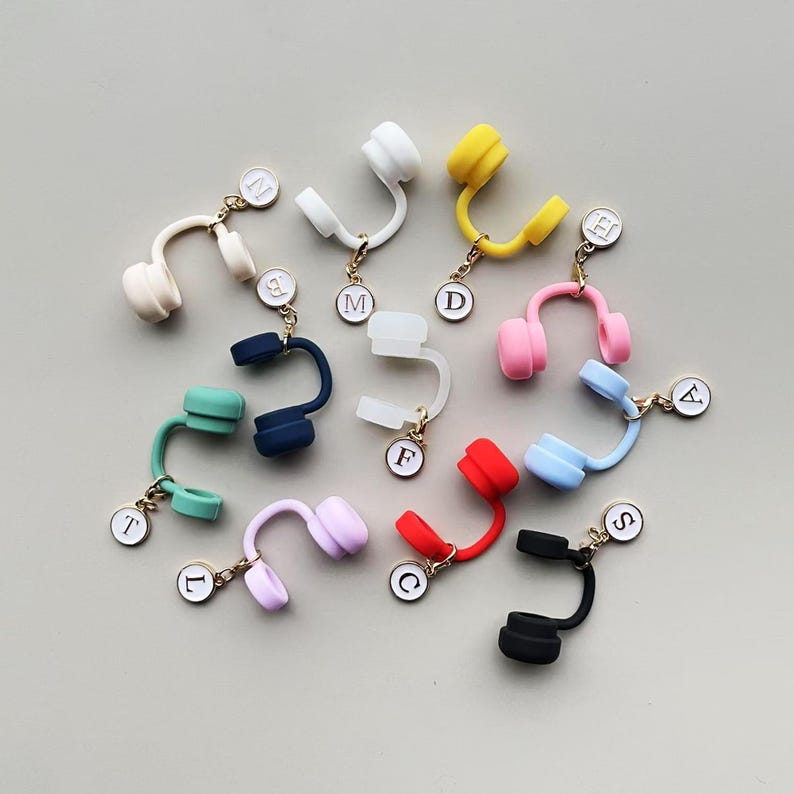 May include: A collection of colorful miniature headphone charms in various colors, including white, yellow, pink, blue, red, green, purple, and black. Each charm has a gold-colored chain and a small, round tag with a letter.