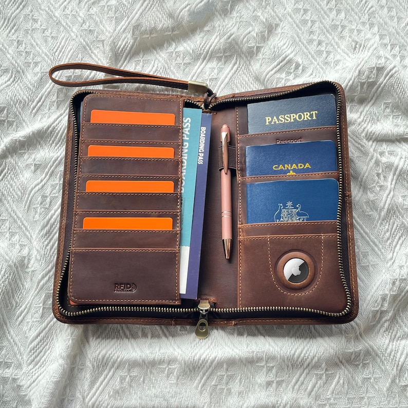 May include: A brown leather travel wallet with a zipper closure, featuring multiple card slots, a pen, and compartments for a passport and boarding pass. The wallet includes an RFID tag and a small circular device.
