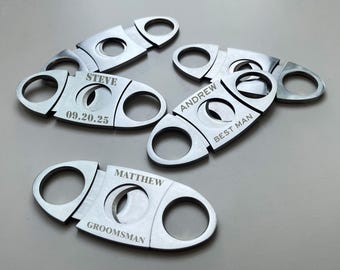 May include: A collection of silver cigar cutters, each with a circular blade and handle. Some cutters are engraved with names and dates, including "STEVE" and "09.20.25", "ANDREW BEST MAN", and "MATTHEW GROOMSMAN". The cutters are arranged on a light gray surface.