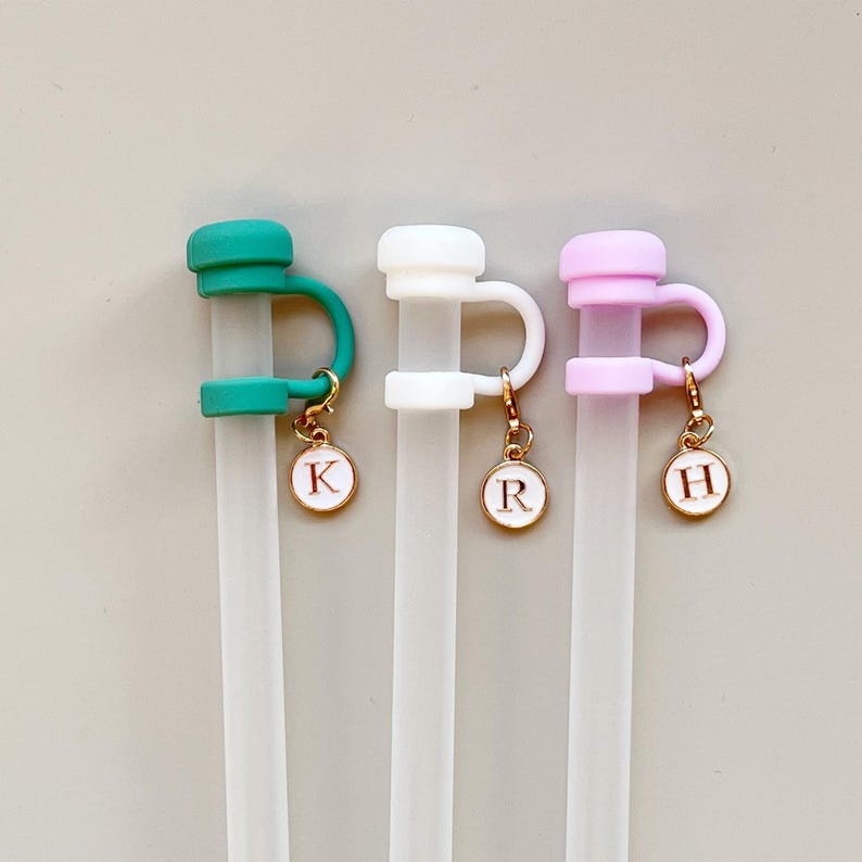 May include: Three reusable clear straws with colorful silicone toppers in teal, white, and pink. Each straw has a gold charm with a white enamel background and a letter: K, R, and H. The charms are attached to the silicone toppers.
