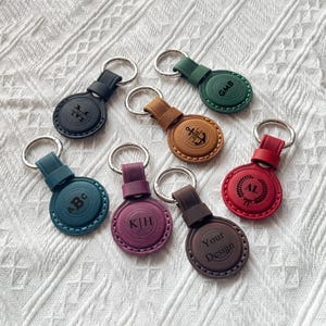 May include: A collection of personalized leather keychains in various colors, including black, green, brown, teal, red, and purple. Each keychain features a silver ring and a circular leather tag with engraved initials, an anchor, or the words "Your Design."