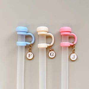 May include: Three reusable straw toppers in pastel colors: light blue, cream, and pink. Each topper has a loop and a charm with a letter: F, G, and J. The charms are gold-colored.