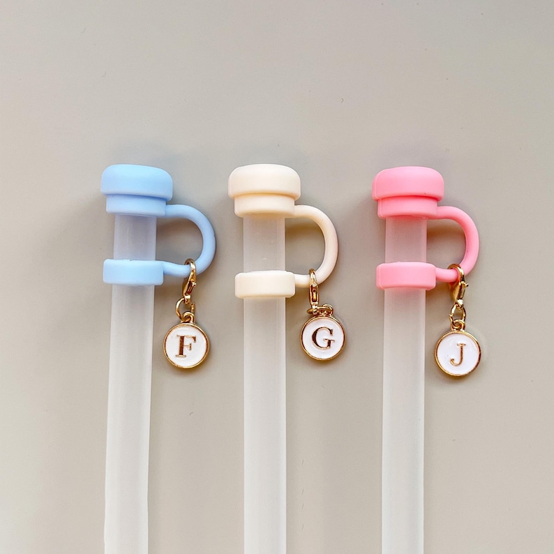 May include: Three reusable straw toppers in pastel colors: light blue, cream, and pink. Each topper has a loop and a charm with a letter: F, G, and J. The charms are gold-colored.