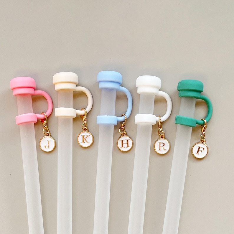 May include: Five reusable clear straws with colorful toppers in pink, cream, light blue, white, and green. Each straw has a matching topper with a gold charm featuring a letter. The letters are J, K, H, R, and F.