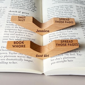 May include: Three wooden bookends shaped like a book page. The bookends are labeled "Spread Those Pages", "Good Girl", and "Jessica".