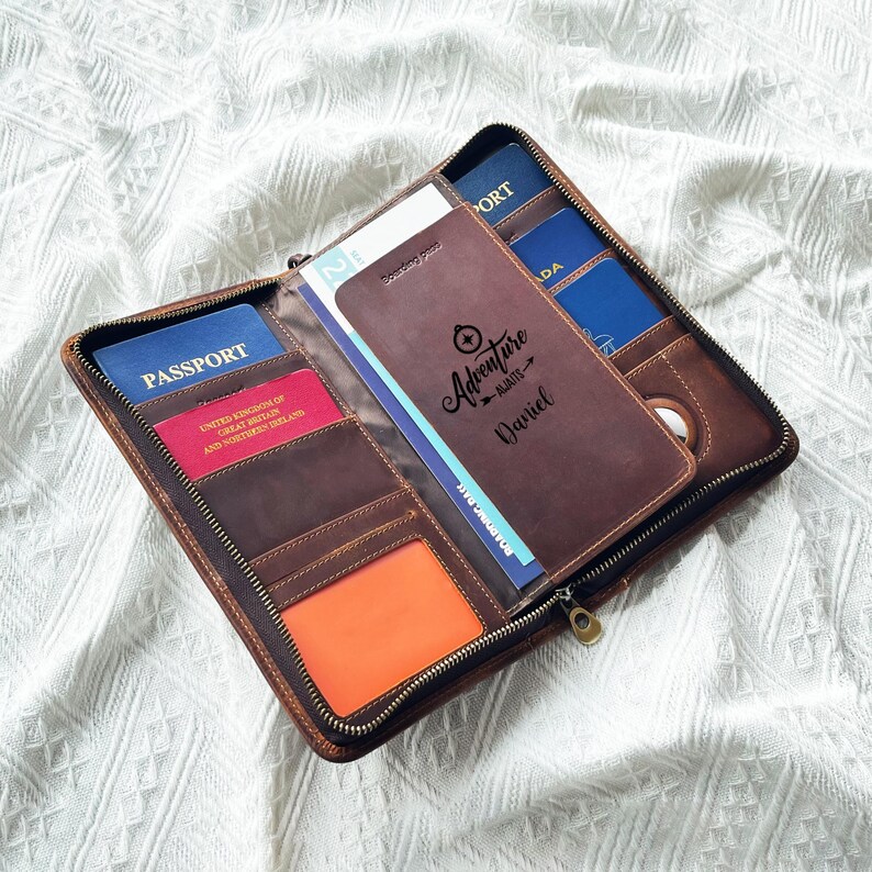 May include: A brown leather travel wallet with a zipper closure, holding passports and cards. The wallet has multiple compartments and features the text "Adventure Awaits Daniel". The wallet is open, displaying the contents.
