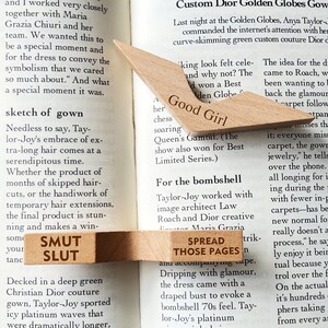 Puede incluir: Marcadores de libros de madera con texto grabado. Un marcador dice "Good Girl" y el otro "Smut Slut" y "Spread Those Pages". Los marcadores son de color marr&oacute;n claro y est&aacute;n colocados sobre un libro.