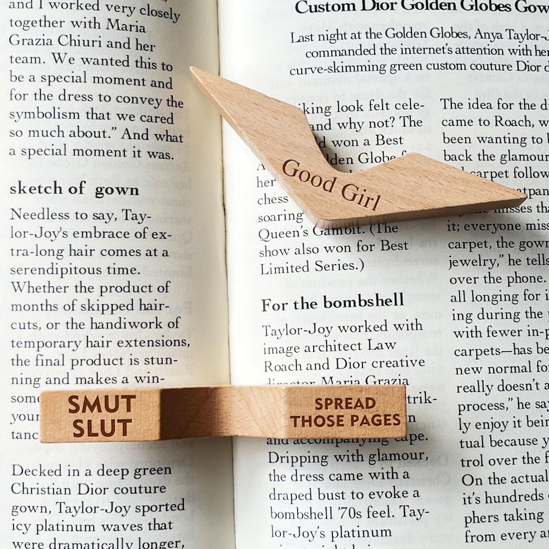 Puede incluir: Marcadores de libros de madera con texto grabado. Un marcador dice "Good Girl" y el otro "Smut Slut" y "Spread Those Pages". Los marcadores son de color marr&oacute;n claro y est&aacute;n colocados sobre un libro.