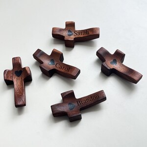 May include: A collection of five wooden crosses, each with a dark brown finish. Each cross features a heart-shaped design and a name engraved on it: Sarah, Carol, and Brandon. The crosses are arranged on a white surface.