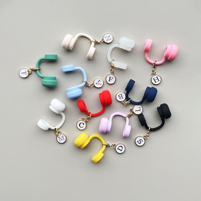 May include: A collection of colorful headphone charms in various hues, including white, pink, green, blue, red, purple, yellow, and black. Each charm has a gold-colored chain and a small, round initial tag. The charms are arranged on a light gray surface.
