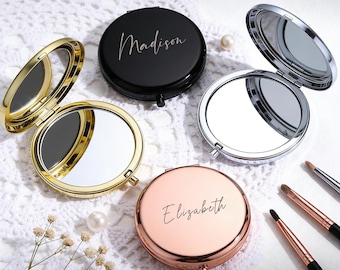 May include: A collection of personalized compact mirrors in gold, black, silver, and rose gold. Each round mirror is open, revealing a reflective surface. The black and rose gold mirrors have names printed on them. Makeup brushes are also present.