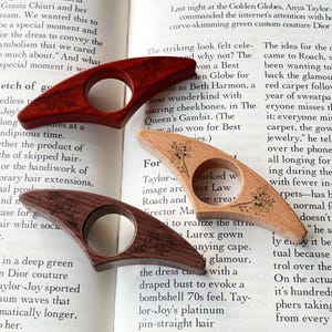 May include: Three wooden thumb page holders in varying shades of brown. Each has a circular opening and a floral design. The page holders are resting on the open pages of a book.
