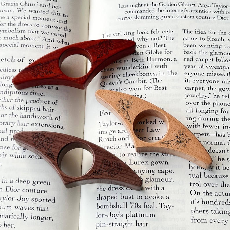 May include: Three wooden thumb page holders in varying shades of brown. Each has a circular opening and a floral design. The page holders are resting on the open pages of a book.