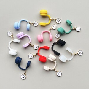 May include: A collection of colorful miniature headphone charms in various colors, including blue, yellow, pink, and red. Each charm has a gold-colored chain and a small, round tag with a letter. The charms are arranged on a light gray surface.