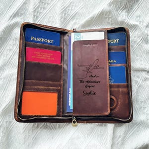 May include: A brown leather travel wallet, open to display passports, boarding passes, and cards. The wallet has multiple compartments and a zipper closure. A personalized passport holder is visible, with the text "And so The Adventure Begins Sophie".