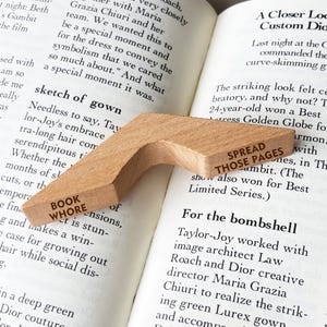 Puede incluir: Un marcap&aacute;ginas de madera con las palabras "BOOK WHORE" y "SPREAD THOSE PAGES" impresas. Descansa sobre las p&aacute;ginas abiertas de un libro.