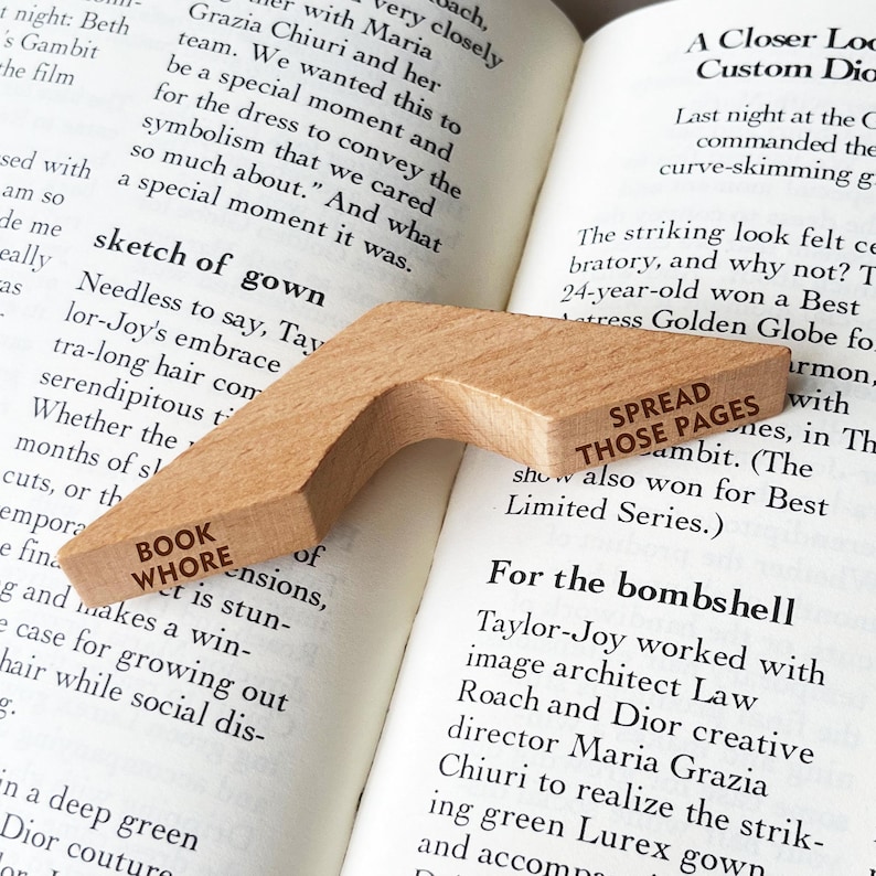 Puede incluir: Un marcap&aacute;ginas de madera con las palabras "BOOK WHORE" y "SPREAD THOSE PAGES" impresas. Descansa sobre las p&aacute;ginas abiertas de un libro.