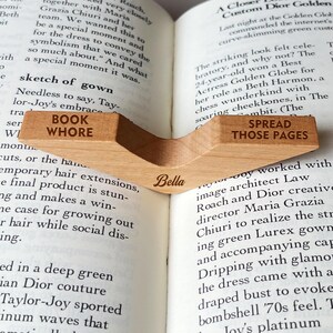 Puede incluir: Un marcap&aacute;ginas de madera clara con las palabras "BOOK WHORE" y "SPREAD THOSE PAGES" grabadas. El marcap&aacute;ginas tiene el nombre "Bella" grabado. Est&aacute; colocado en un libro abierto.
