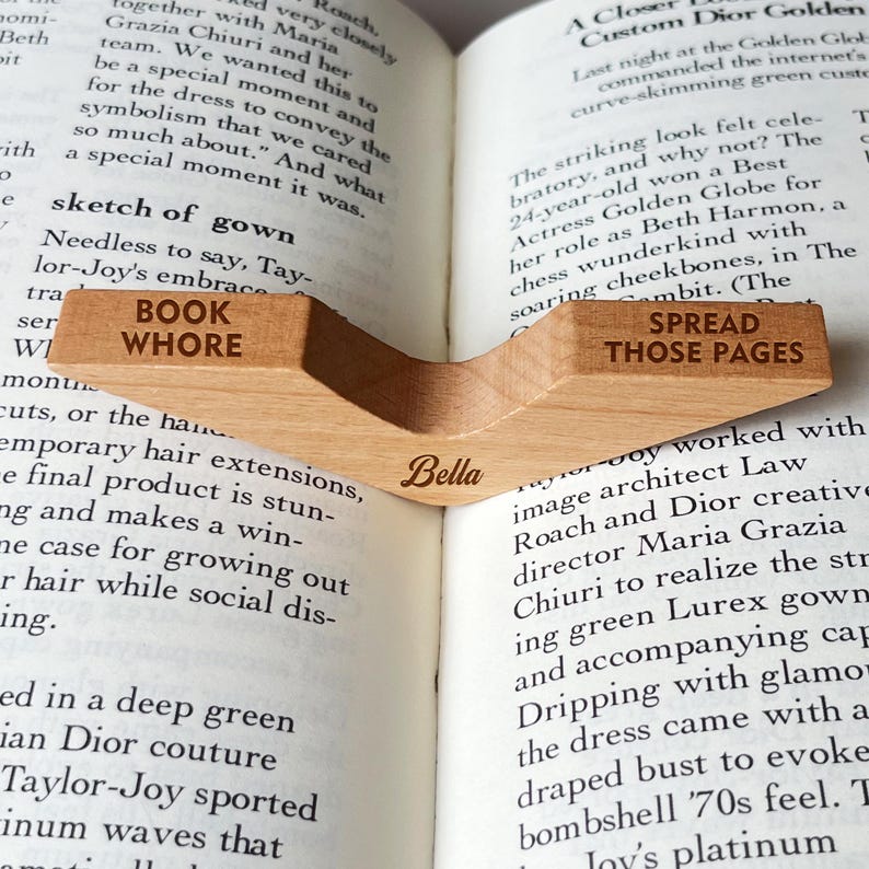 Puede incluir: Un marcap&aacute;ginas de madera clara con las palabras "BOOK WHORE" y "SPREAD THOSE PAGES" grabadas. El marcap&aacute;ginas tiene el nombre "Bella" grabado. Est&aacute; colocado en un libro abierto.