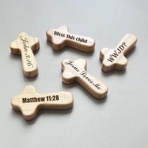 May include: Five wooden crosses with engraved text. The crosses are light brown and feature various inscriptions, including "John 3:16", "Bless This child", "WWJD?", "Jesus Loves Me", and "Matthew 11:28".