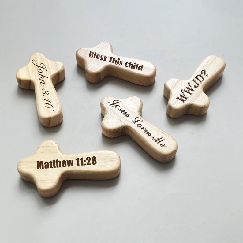 May include: Five wooden crosses with engraved text. The crosses are light brown and feature various inscriptions, including "John 3:16", "Bless This child", "WWJD?", "Jesus Loves Me", and "Matthew 11:28".