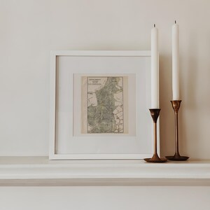 May include: A framed map print with a white border, displayed on a white surface. Two white candles stand tall in brass candle holders. The map features green and brown tones, adding a vintage aesthetic to the decor.
