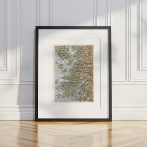 May include: A framed vintage map print featuring detailed topographical lines, geographical labels, and muted earth tones of tan, green, and blue. The map is encased in a simple black frame with a white mount, displaying a vertical orientation.