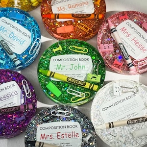 May include: A selection of round resin paperweights, each with a glittery finish. The paperweights feature a miniature composition book design, a crayon, and other stationery items. Each paperweight is a different colour, including yellow, blue, orange, pink, purple, green, and silver.