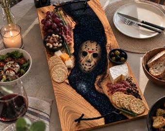 May include: A rectangular wooden serving board with a central river of dark blue glittery resin featuring an embedded hockey mask. The board has black branch-shaped handles and is arranged with grapes, nuts, crackers, cheese, and cured meats.