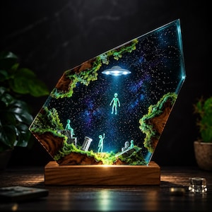 May include: A trapezoidal resin lamp featuring a deep space scene with a glowing UFO hovering above small alien figures. The diorama includes textured green moss, dark wood elements, and a starry night sky background set on a rectangular wood base.