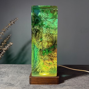 May include: A rectangular lamp with a wooden base. The lamp's body is a clear resin filled with green and yellow organic shapes. The lamp is lit from the bottom, creating a glowing effect. A white cord extends from the base.